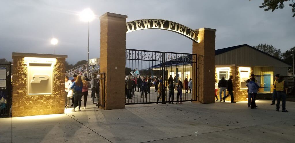 Entrance to Dewey Field at Perry Community School District, featuring a lit archway, ticket booths, and a crowd entering the stadium.
