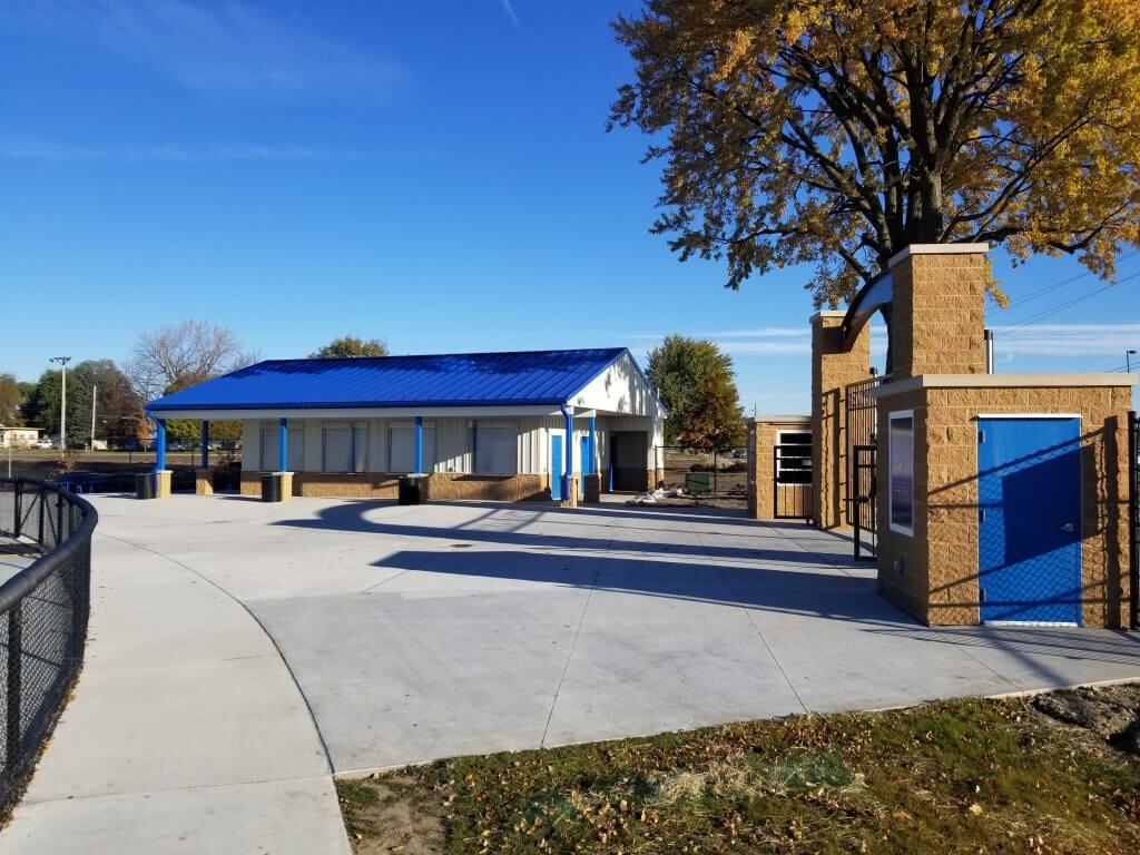 New concession and restroom building with blue roof at Perry Community School District athletic facility, featuring adjacent ticket booths and stadium entrance under a clear sky.