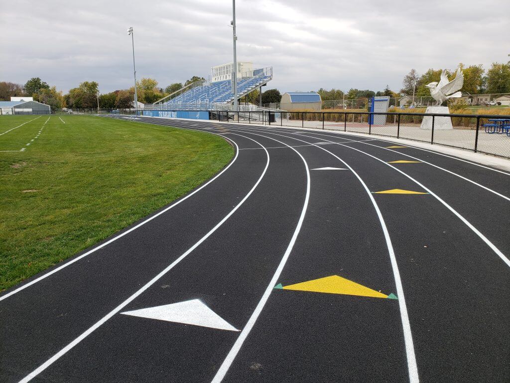 Running track with lane markings and directional arrows, adjacent to a grassy field and blue bleachers, under a cloudy sky.