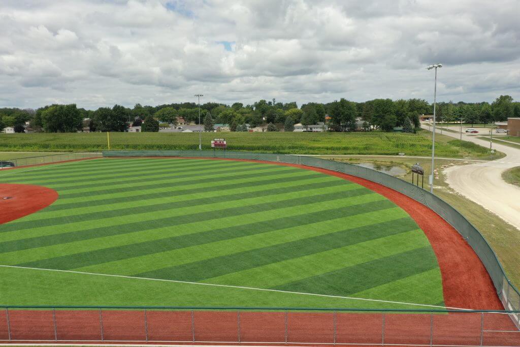 Baseball field with a striped turf outfield and red dirt warning track, surrounded by a fence, under a cloudy sky.