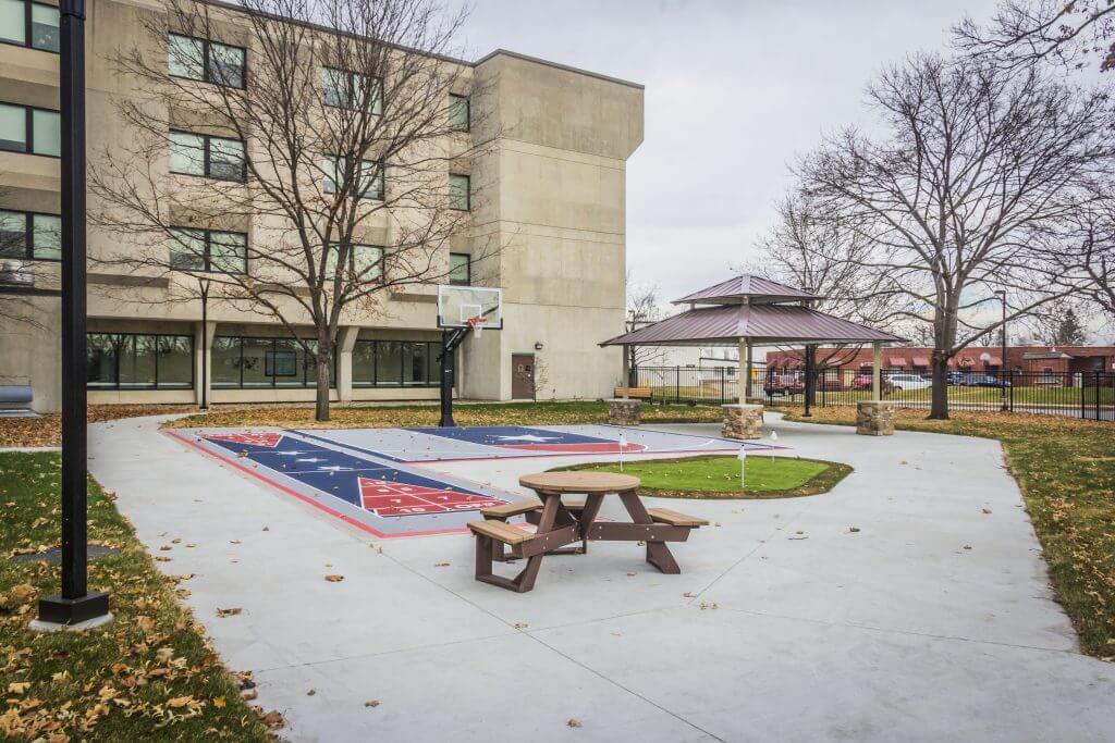 Courtyard of the Iowa Veterans Home featuring a basketball court, picnic table, and gazebo, surrounded by trees and a multi-story building.