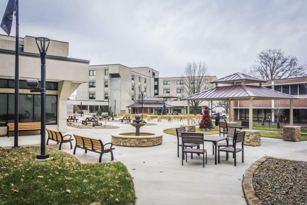 Courtyard at the Iowa Veterans Home featuring a central fountain, benches, tables, a gazebo, and surrounding buildings, with overcast skies.