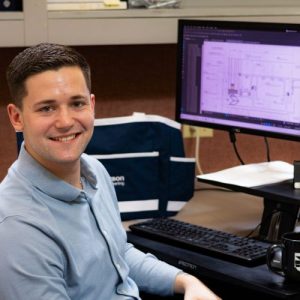 Engineer at a desk smiling, with a computer screen displaying technical drawings and a bag labeled "Larson Engineering" in the background.