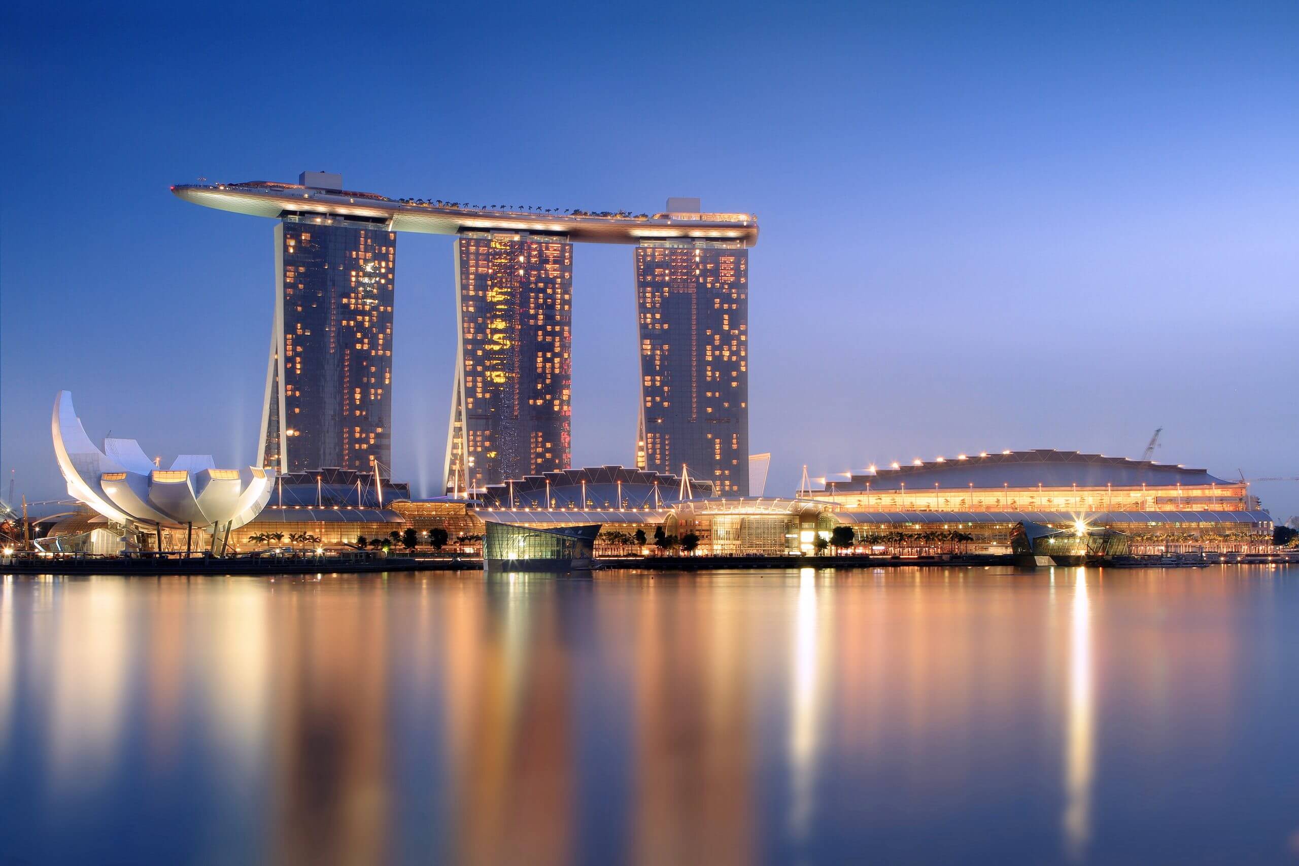 Marina Bay Sands complex with its iconic three towers and sky park, illuminated against a twilight sky, reflecting on the calm waters below.
